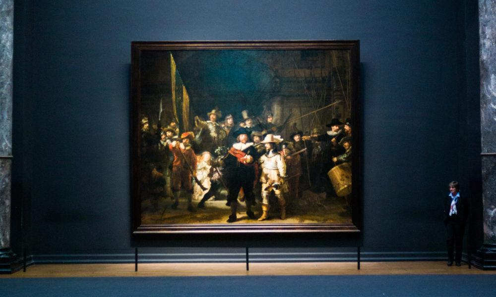 Amsterdamliebe Rijksmuseumstour Nachtwache von Rembrandt hinter dunkelblauer Wand