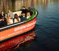 A group of tourists on a boat tour in Amsterdam. The boat has an orange and white color scheme with green accents.
