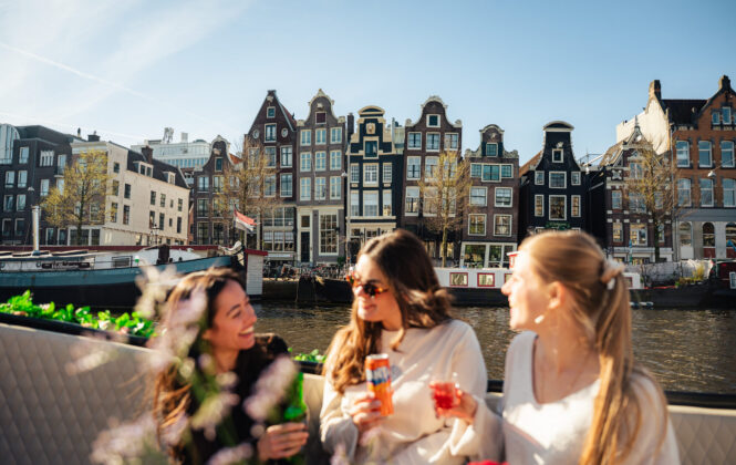 Three women on a boat in Amsterdam, smiling and holding drinks. They appear to be on a city trip enjoying a sunny day.