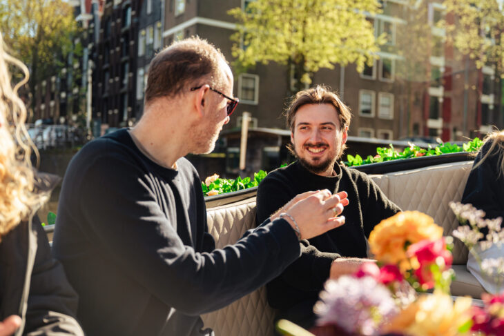 Two men chatting and smiling on an outdoor patio. They appear to be on a coffee break.
