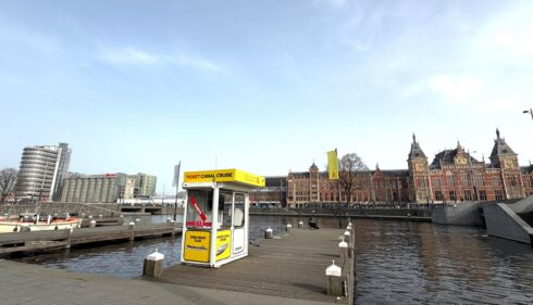 A small yellow and white kiosk by a canal in Amsterdam. The historic buildings and modern architecture are visible in the background.