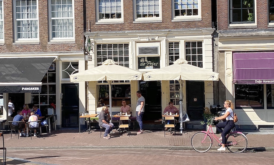 Fahrradfahrerin fährt vor sitzenden Menschen vor dem Café de Prins vorbei