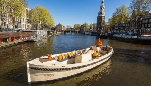 A white boat on a canal in Amsterdam. The boat has orange and yellow life jackets on board.