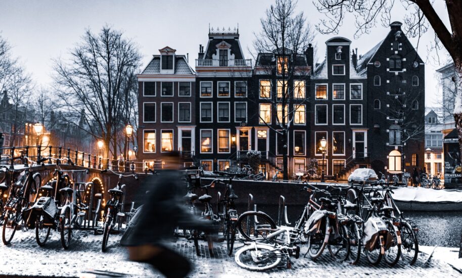 A snowy scene of a canal in Amsterdam with many bicycles parked along the side and traditional Dutch buildings in the background. The buildings are lit up warmly in the evening.