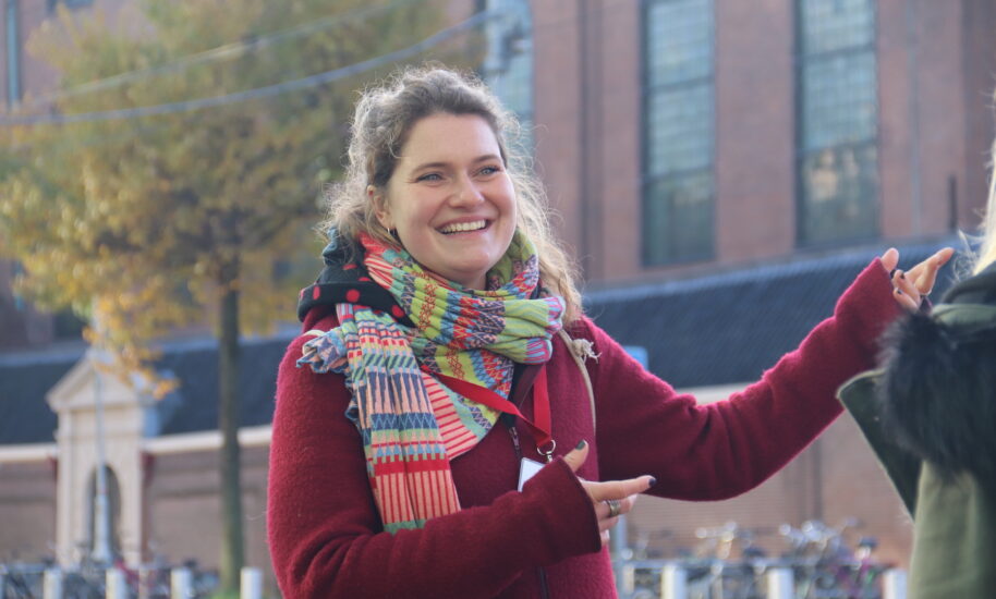 A smiling woman wearing a colorful scarf. She is standing outside in a urban setting.