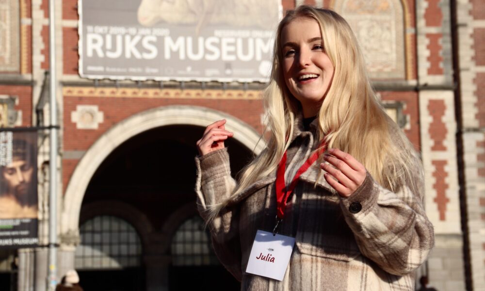 Blonde woman in beige jacket standing outside museum. She is wearing a red lanyard around her neck.