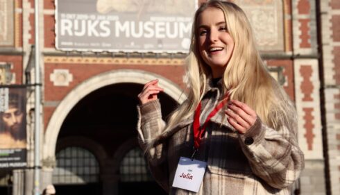 Blonde woman in beige jacket standing outside museum. She is wearing a red lanyard around her neck.