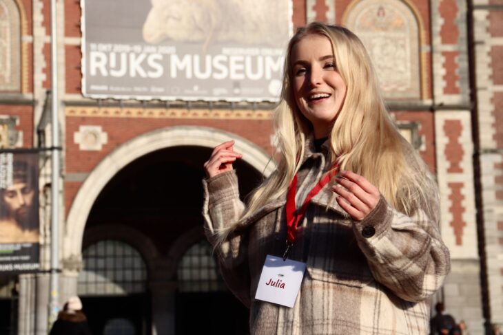 Blonde woman in beige jacket standing outside museum. She is wearing a red lanyard around her neck.