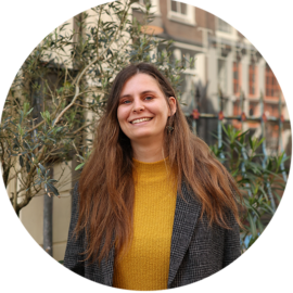 A smiling woman with long brown hair, wearing a yellow sweater and a grey blazer. She appears to be in a city setting.