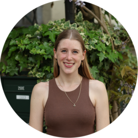 A smiling woman with long brown hair, wearing a brown tank top and a necklace. She is standing in front of green plants.