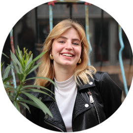 A young woman with blonde hair smiling. She is wearing a white shirt and a black leather jacket.