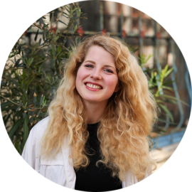 A young woman with long blonde hair smiling. She is standing in front of some plants.