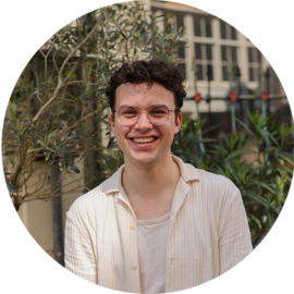 A smiling young man standing outside with plants behind him. He is wearing a white shirt.