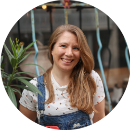 A smiling woman with long hair wearing dungarees. She is outdoors.