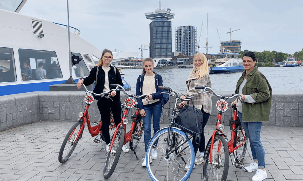 Group of friends posing with bicycles in Amsterdam. They are standing on a dock with a boat and cityscape in the background.
