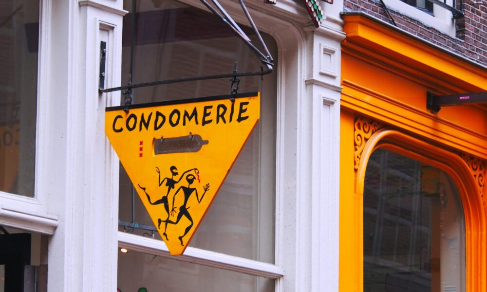 A yellow triangular sign reading Condomerie hangs outside a store. A display of bottles is visible through the window.