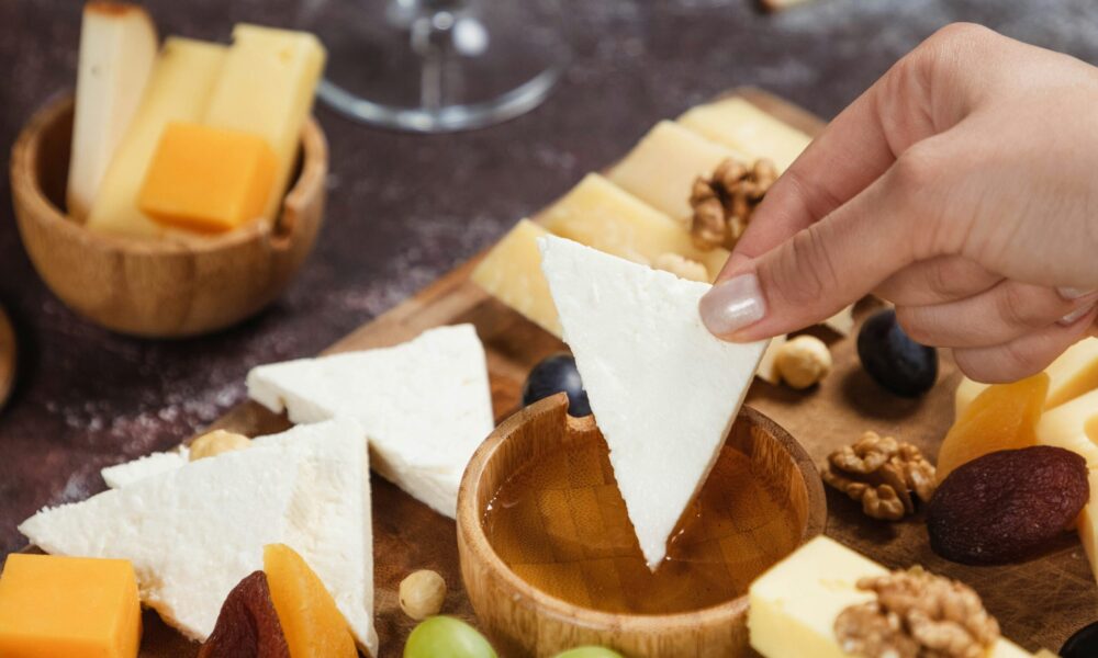 A cheese platter with a variety of cheeses and fruits. A hand picking up a slice of white cheese.