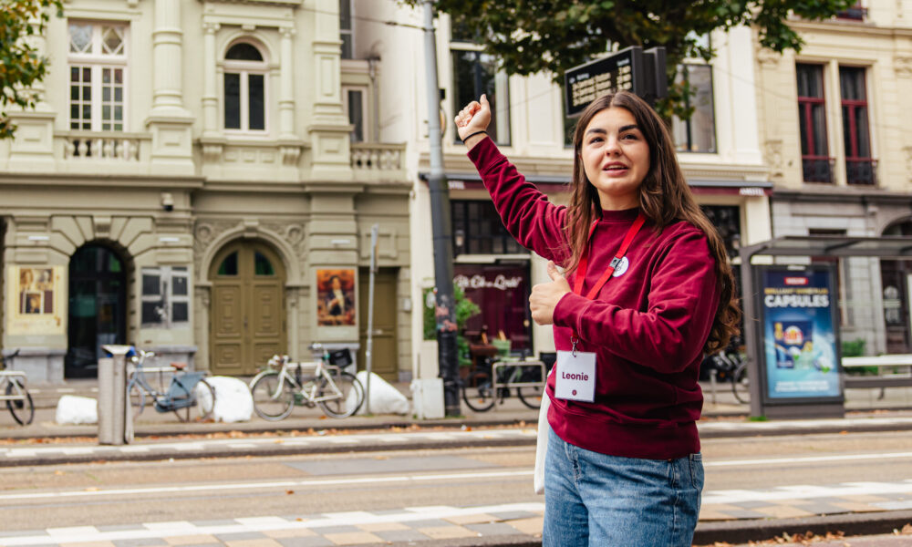 A smiling tour guide in a red sweater pointing at a street in Amsterdam. She is wearing a lanyard with a badge.