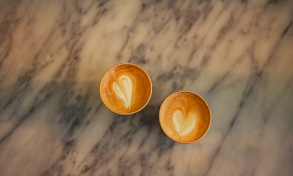 Two cups of coffee on a marble table. The coffee cups have heart designs on top.
