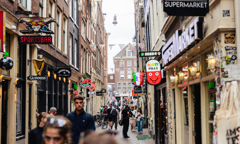 People walking down a narrow shopping street in Amsterdam. Colorful signs hang above the shops.