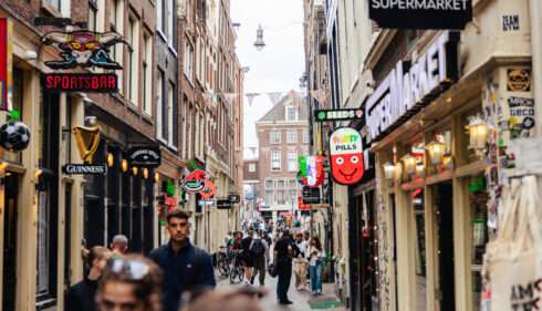 People walking down a narrow shopping street in Amsterdam. Colorful signs hang above the shops.