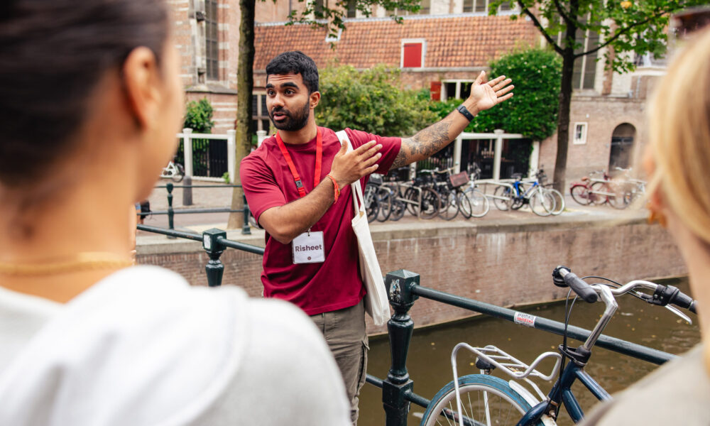A tour guide explaining Amsterdam city to tourists. He is standing by a canal with bicycles.