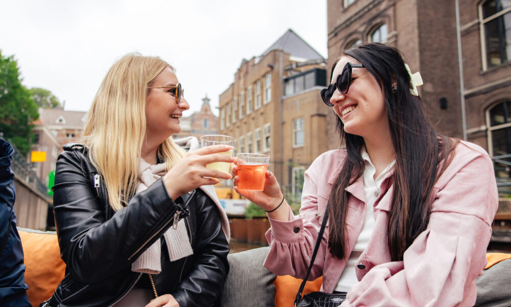 Two women smiling and toasting with drinks on a terrace in Amsterdam. They are enjoying a sunny day.