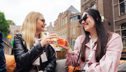 Two women smiling and toasting with drinks on a terrace in Amsterdam. They are enjoying a sunny day.