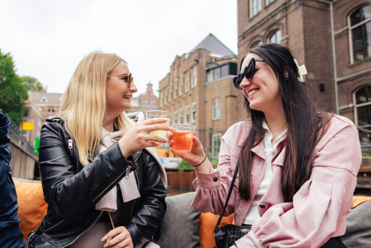 Two women smiling and toasting with drinks on a terrace in Amsterdam. They are enjoying a sunny day.