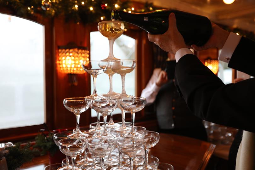 A waiter pouring champagne into glasses on a train. The glasses are stacked in a tower shape.