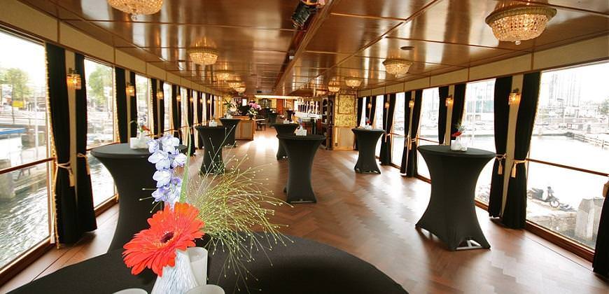 A stylish event room with black tables and flowers. Large windows offer views of boats and the city.