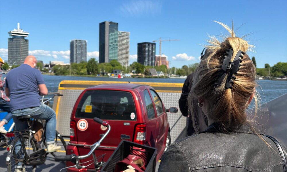 People on a ferry with bikes and a car. A woman with her back to the camera looks out at Amsterdam.