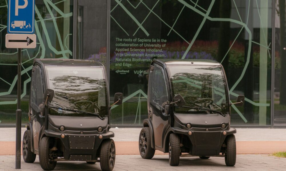 Two small black electric cars parked on a sidewalk. They are next to a parking sign.