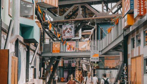 People walking through a vibrant street market in Amsterdam. Colorful signs and buildings line the walkway.