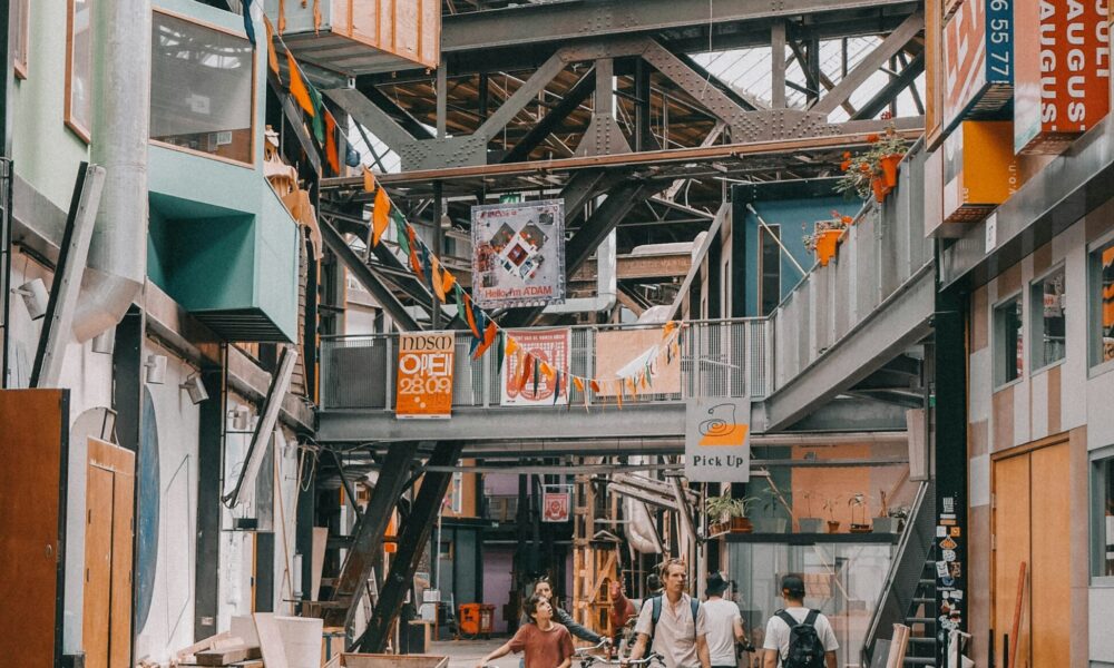 People walking through a large industrial space with metal beams and colorful signs.
