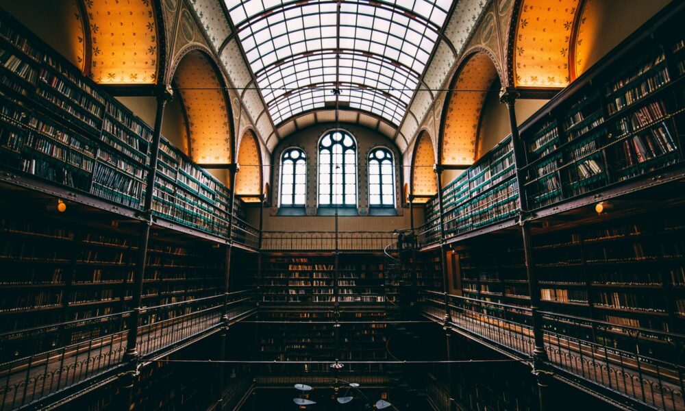 A dimly lit library with a high ceiling and bookshelves. The room has a glass roof and large windows.