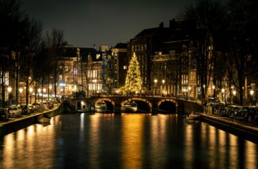 A canal in Amsterdam at night with a Christmas tree in the background. The water reflects the lights of the buildings and street lamps.