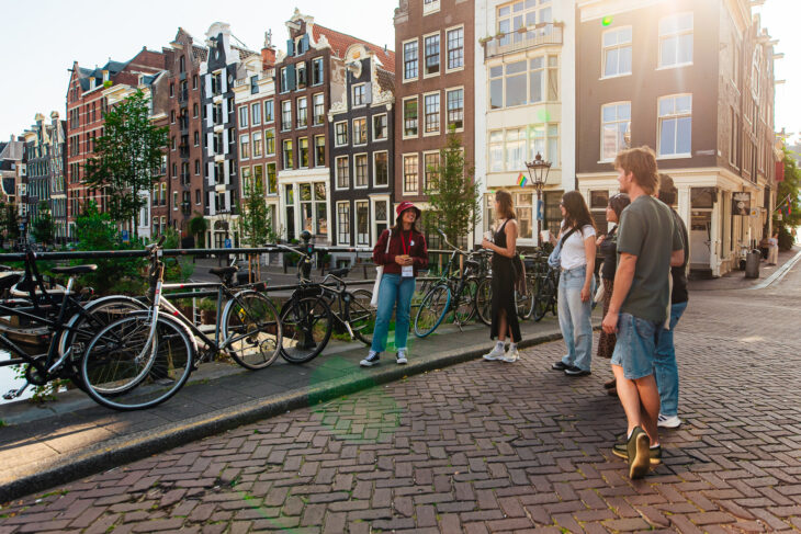 Group of people on a brick sidewalk next to bicycles on a canal street in Amsterdam. The background features historic buildings along the water.