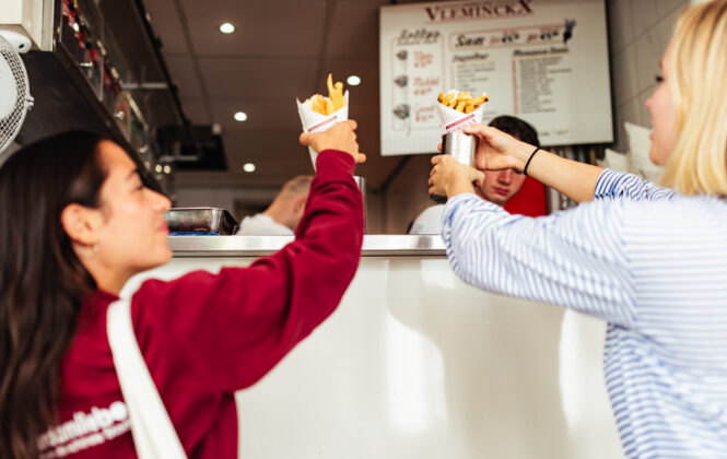 A tour guide and tourist on a food tour in Amsterdam. They are both grabbing a cone of french fries from a stall.
