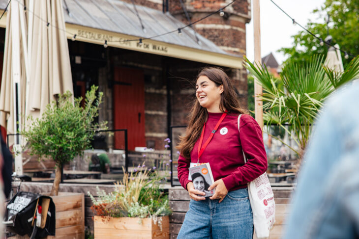 Tour Guide Leonie with a red sweater and blue jeans holding a guidebook. She is standing in front of a cozy cafe in Amsterdam.