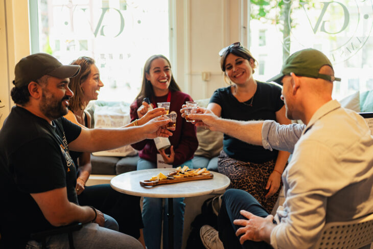 A group of tourists enjoy liquor and cheese on a food tour through Amsterdam. There is a cheese board on the small round table between them. They are all smiling and enjoying themselves