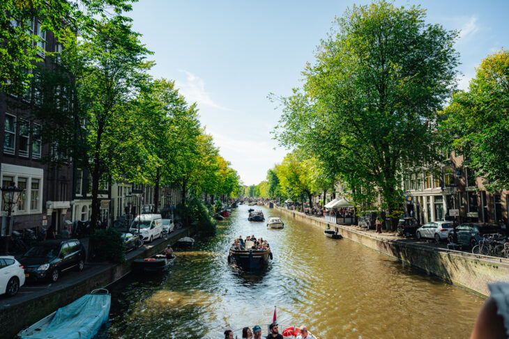 A canal in Amsterdam with boats and green trees. The waterway reflects the sunny day.