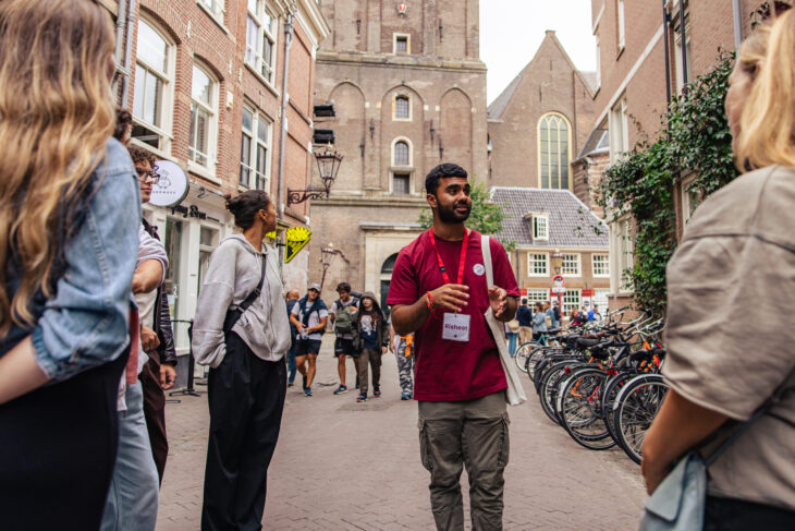 A tour guide leading a group of tourists through Amsterdam. The tour guide is wearing a name tag with the name Risheet on it.
