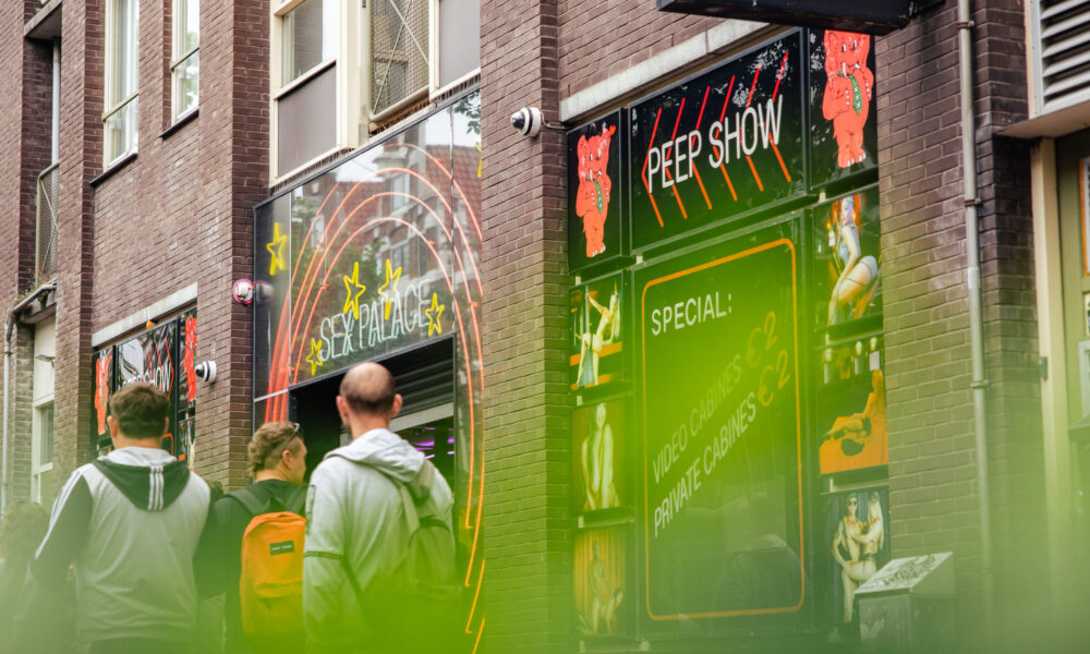People walking past a colorful entertainment venue in Amsterdam while on a Red Light District tour. A sign reads 'PEEP SHOW'.
