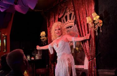 Drag Queen show café the queens head. Blonde performer in a white dress on stage. The performer appears to be in a theatrical setting.