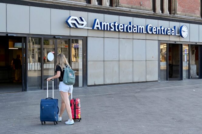 A woman with luggage walking towards Amsterdam Centraal Station. The station sign is visible in the background.