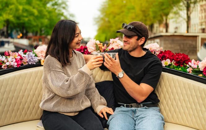 A young couple enjoys a romantic moment on a boat, surrounded by flowers. They are smiling and holding drinks.